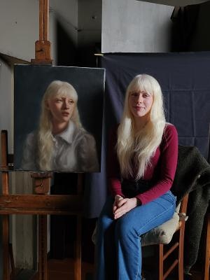 A fair-haired sitter seated beside her completed portrait on an easel